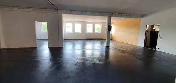 Große, helle Halle mit gleichmäßig beschichtetem dunklem Boden und Fensterfront.
